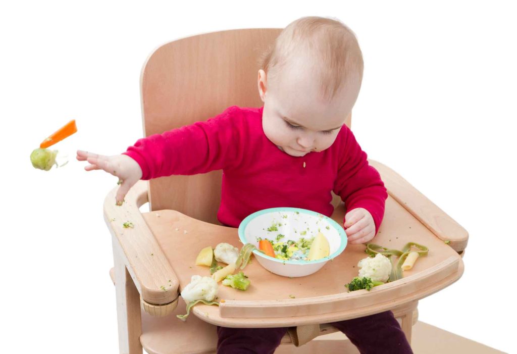 Ребенок сидит в детском стуле и разбрасывает вареные овощи
